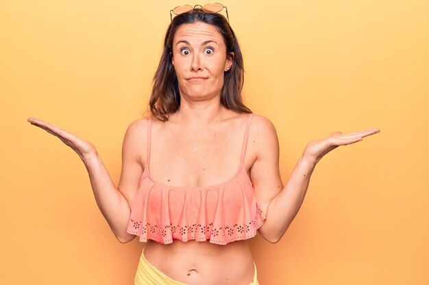 Young beautiful brunette woman wearing bikini standing over isolated yellow background clueless and confused with open arms no idea and doubtful face