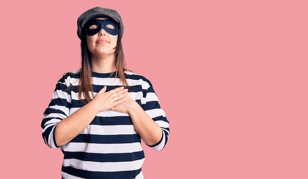 Free photo young beautiful brunette woman wearing burglar mask smiling with hands on chest with closed eyes and grateful gesture on face. health concept.