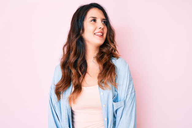 Young beautiful brunette woman wearing casual clothes over pink background looking away to side with smile on face, natural expression. laughing confident.