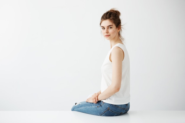 Young beautiful brunette woman with bun smiling sitting over white background.