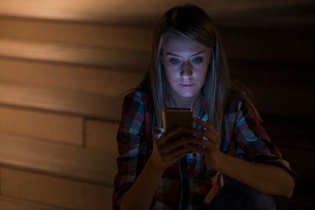 Young beautiful business woman sitting at night lights with mobile smart phone. Evening woman working remotely. Surfing internet courses, reading books, watching videos online, texting message.
