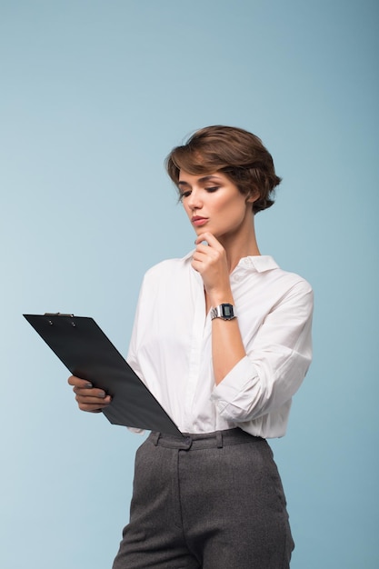 Молодая красивая деловая женщина с темными короткими волосами в белой рубашке задумчиво смотрит на папку с документами на синем изолированном фоне