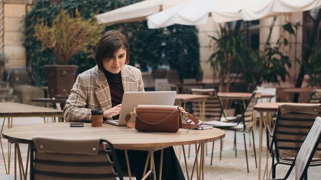 Young beautiful businesswoman discussing work issues with colleagues by video call using laptop during coffee break in courtyard of cafe