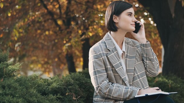 도시 거리 현대 기술에서 일하는 동안 스마트폰으로 말하는 격자 무늬 블레이저를 입은 젊은 아름다운 여성 사업가