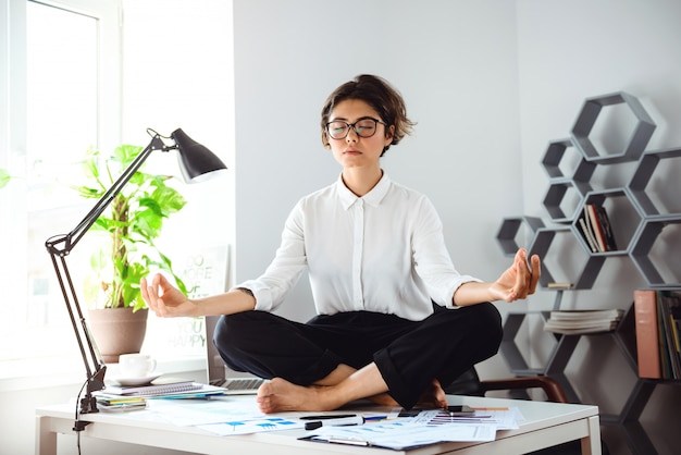 Free photo young beautiful businesswoman meditating on table at workplace in office.