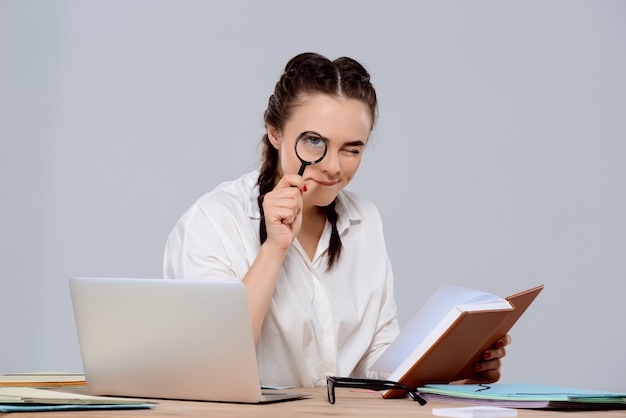 Young beautiful businesswoman sitting at workplace, holding book and magnifier