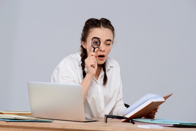 Young beautiful businesswoman sitting at workplace, holding book and magnifier