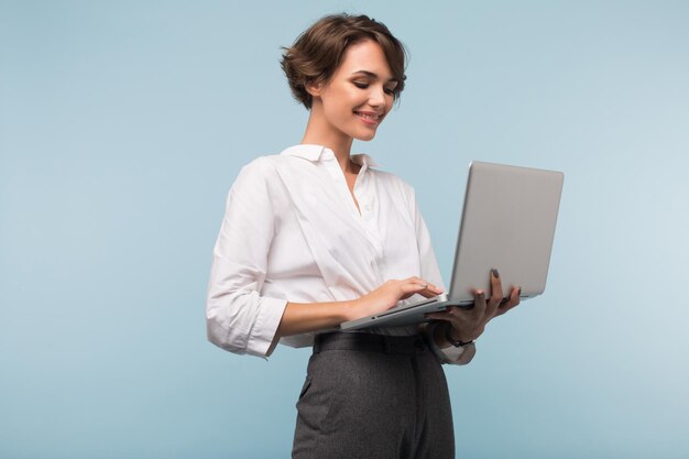 Young beautiful businesswoman with dark short hair in white shirt happily working on laptop over blue background isolated