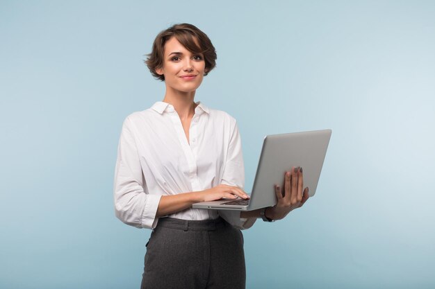 Молодая красивая деловая женщина с темными короткими волосами в белой рубашке держит ноутбук в руках, мечтательно глядя в камеру на синем фоне