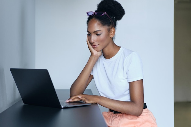 Молодая красивая случайная афроамериканка мечтательно работает на ноутбуке в современном офисе