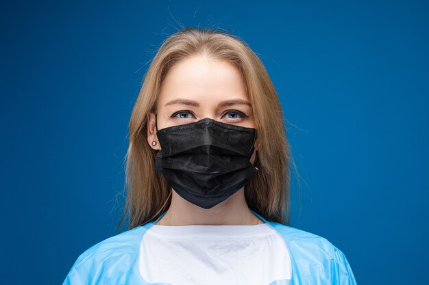 Young beautiful caucasian female in blue medical gown and with white medical mask on her face looks on the camera