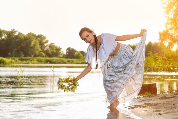 Молодая красивая кавказская женщина, стоящая на берегу реки. Традиционная сельская фотография с девушкой на переднем плане и копировальным пространством. Брюнетка держит венок. Солнечная вспышка