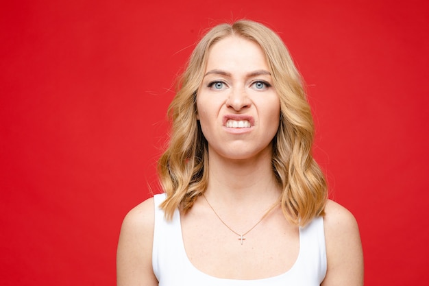 Free photo young beautiful caucasian woman with medium fair wavy hair and nude makeup in white t-shirt looks in disgust, picture isolated on red background