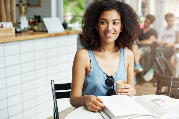 Young beautiful cheerful african woman student smiling laughing sitting in cafe. Books magazines lying on table. Learning and education.