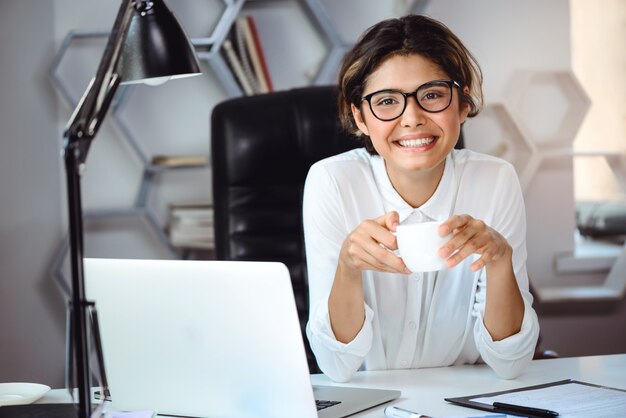 Young beautiful cheerful businesswoman drinking coffee, smiling at workplace in office.