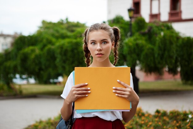Young beautiful cheerful female student, holding folders outdoors
