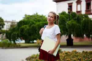 Free photo young beautiful cheerful female student smiling, holding folders outdoors