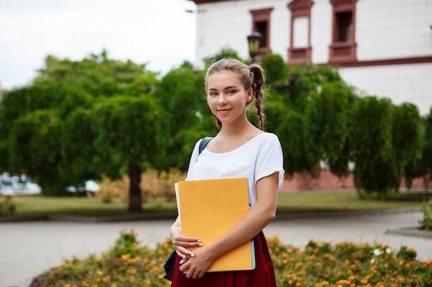 Young beautiful cheerful female student smiling, holding folders outdoors