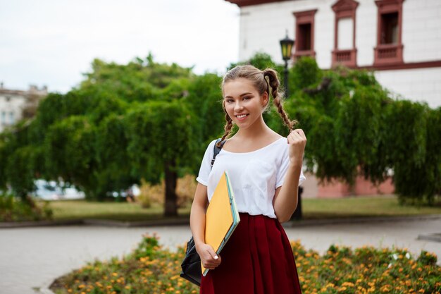 Young beautiful cheerful female student smiling, holding folders outdoors