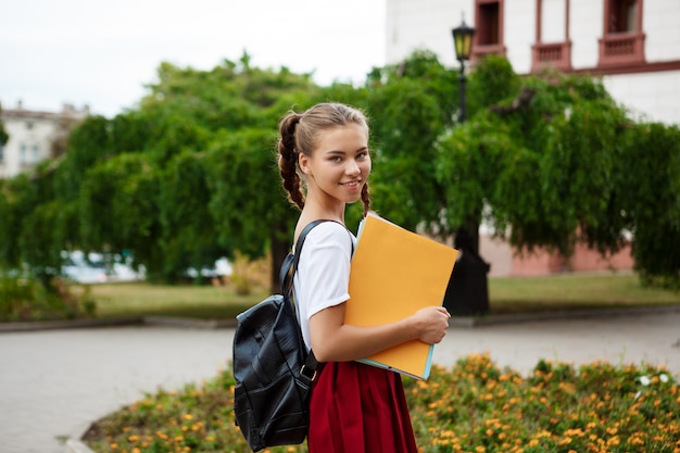 Young beautiful cheerful female student smiling, holding folders outdoors