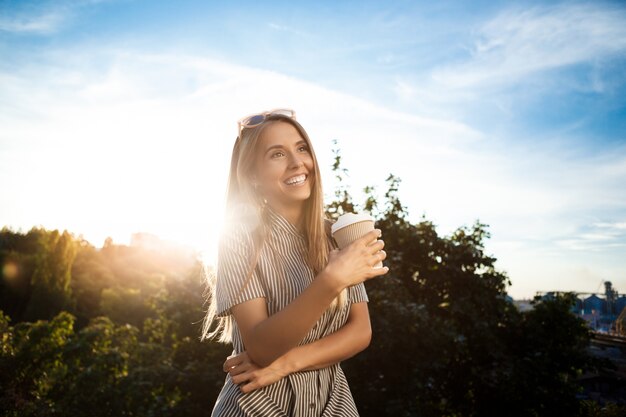 街を歩いて、笑みを浮かべて、コーヒーを保持している若い美しい陽気な女性。