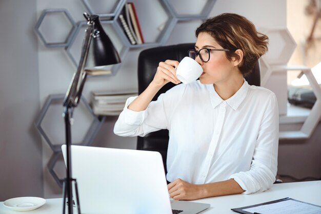 若い美しい自信を持って女性実業家のオフィスの職場でコーヒーを飲みます。