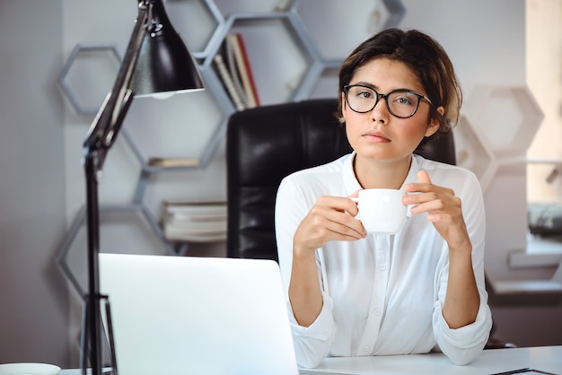 若い美しい自信を持って女性実業家のオフィスの職場でコーヒーを飲みます。