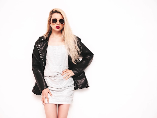 Young beautiful confident female in trendy summer evening silver dress Sexy carefree blond woman posing near white wall in studio Model in black leather biker jacket and red lips