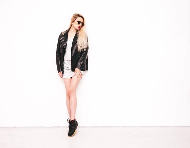 Young beautiful confident female in trendy summer evening silver dress Sexy carefree blond woman posing near white wall in studio Model in black leather biker jacket and sunglasses