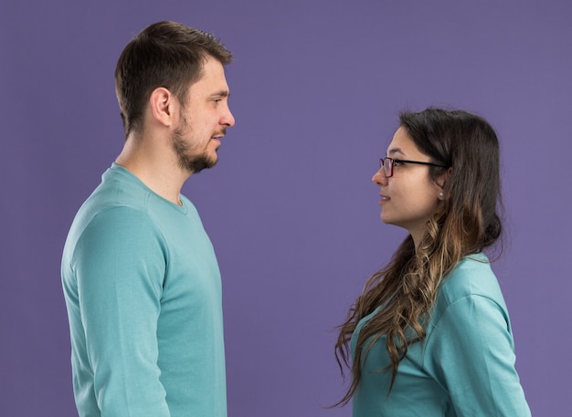 Young beautiful couple in blue casual clothes happy and cheerful man and woman looking at each other happy in love standing over purple wall