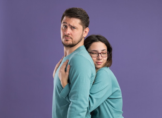 Young beautiful couple in blue casual clothes happy woman hugging her boyfriend happy in love celebrating valentines day standing over purple wall
