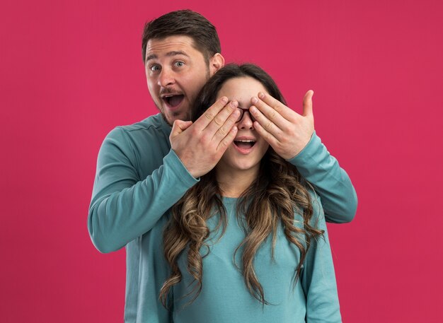 Young beautiful couple in blue casual clothes man closing eyes of his girlfriend making a surprise happy in love together standing over pink wall