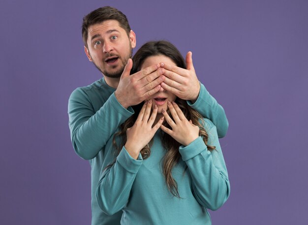 Young beautiful couple in blue casual clothes man closing eyes of his girlfriend making a surprise happy in love together standing over purple wall