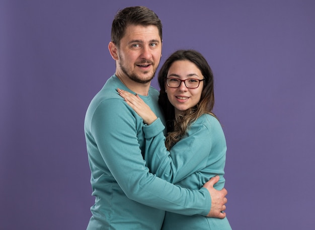 Молодая красивая пара в синей повседневной одежде мужчина и женщина обнимаются, весело улыбаясь, счастливы в любви, вместе стоя над фиолетовой стеной