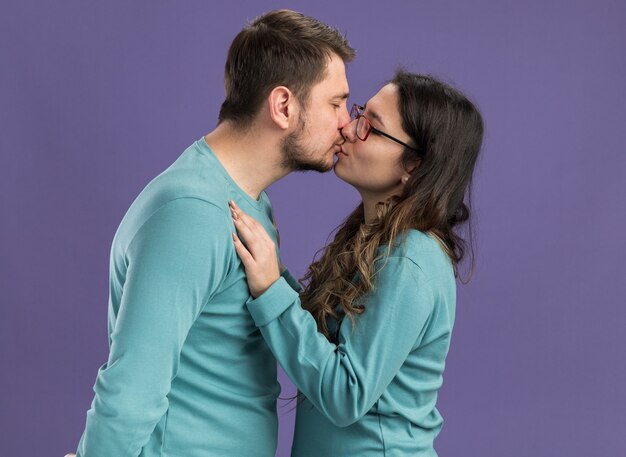 青いカジュアルな服を着た若い美しいカップルの男女が紫の壁の上に立って恋に幸せなキス