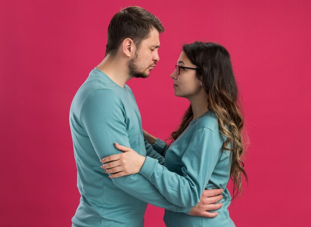青いカジュアルな服を着た若い美しいカップル 男性と女性がお互いを見て、ピンクの壁の上に立って一緒に恋に幸せな彼のガールフレンドにキスをする