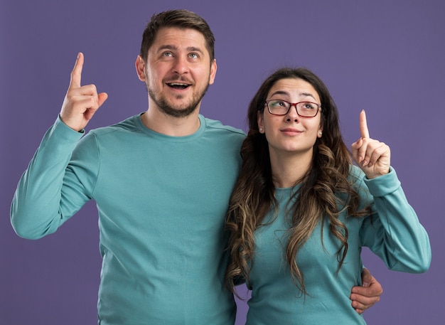 紫色の壁の上に人差し指が立っていることを示すスマートな顔に笑顔で見上げる青いカジュアルな服の男女の若い美しいカップル