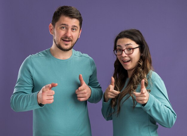 Young beautiful couple in blue casual clothes man and woman  smiling cheerfully pointing with index fingers  happy in love standing over purple wall