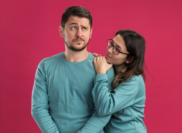 Молодая красивая пара в синей повседневной одежде женщина смотрит с любовью на своего смущенного парня, стоящего над розовой стеной