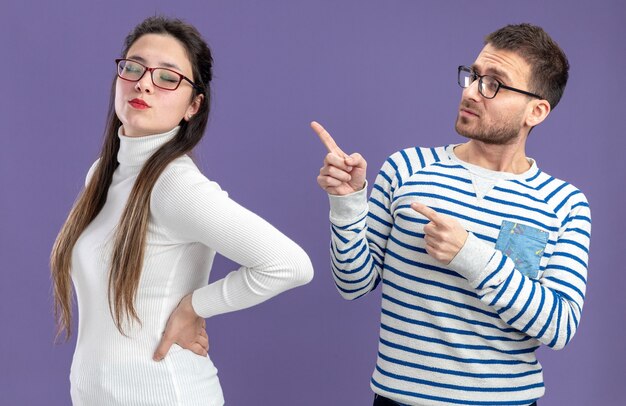 молодая красивая пара в повседневной одежде смущает человека, указывая указательным пальцем на свою серьезную подругу, концепцию Дня святого Валентина, стоящую на фиолетовом фоне