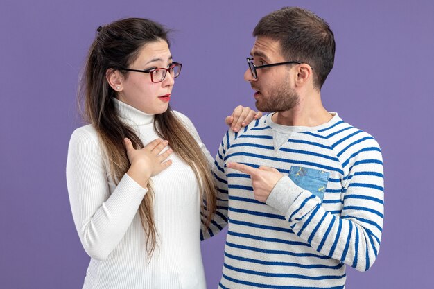 young beautiful couple in casual clothes confused man pointing with index finger at his surprised girlfriend celebrating valentines day standing over purple wall