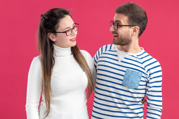 young beautiful couple in casual clothes happy man and woman wearing glasses looking at each other smiling cheerfully celebrating valentines day standing over pink wall