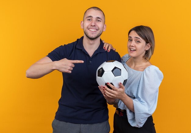 Young beautiful couple in casual clothes smiling woman holding soccer ball while her smiling boyfriend pointing with index finger at ball standing over orange wall