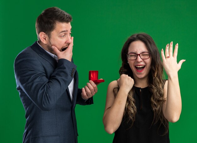 Young beautiful couple confused man holding red box making a proposal to his lovely excited girlfriend showing engagement ring on her finger celebrating valentines day standing over green wall