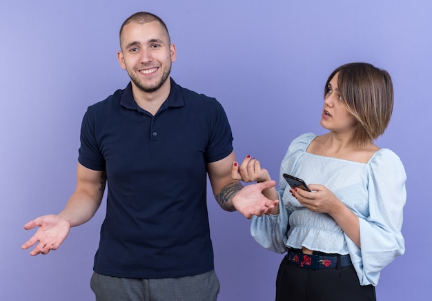 Young beautiful couple confused woman with smartphone with arm raised in indignation looking at her careless smiling boyfriend standing over blue wall