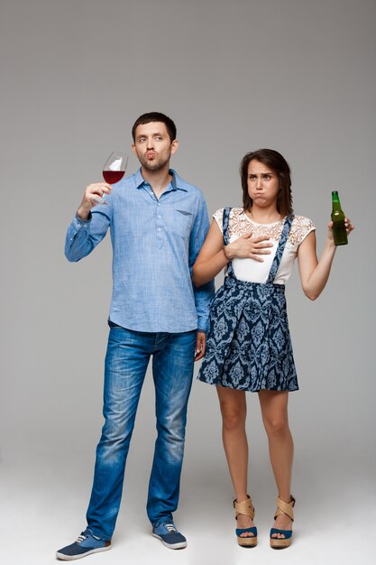 Young beautiful couple drinking wine and beer over grey wall