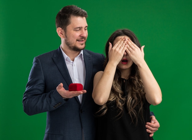 Free photo young beautiful couple handsome man going to make a proposal to his lovely excited girlfriend with closed eyes celebrating valentines day standing over green wall