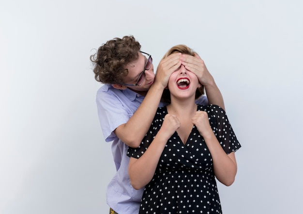 Uomo felice delle giovani belle coppie che copre gli occhi alla sua ragazza allegra che fa sorpreso sul muro bianco