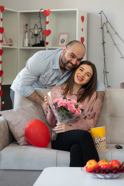 Young beautiful couple happy man giving a bouquet of flowers to his smiling girlfriend sitting on a couch in light living room celebrating international women's day