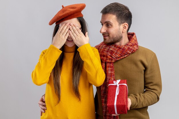 young beautiful couple happy man giving a present for his smiling girlfriend in beret who covering eyes with hands happy in love together celebrating valentines day standing over white background